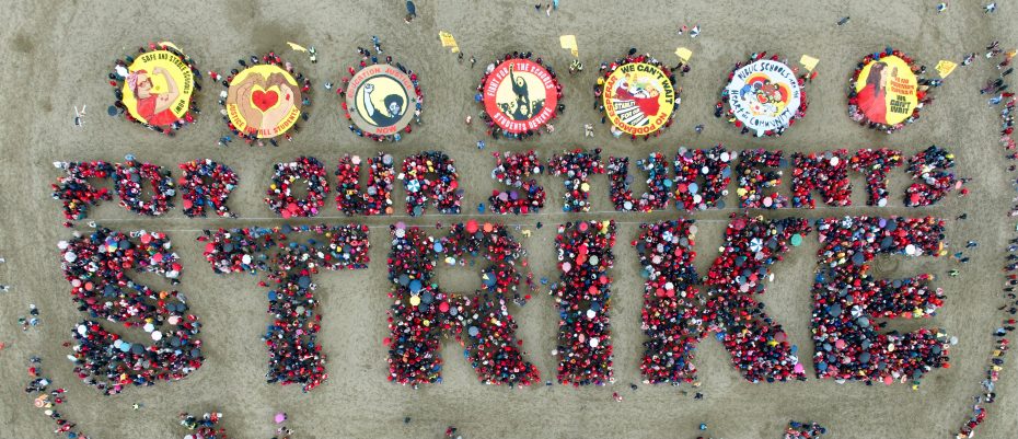 Aerial view of a teachers strike, with a large crowd forming the words "FOR OUR STUDENTS STRIKE" and circular banners displayed above on sandy ground.