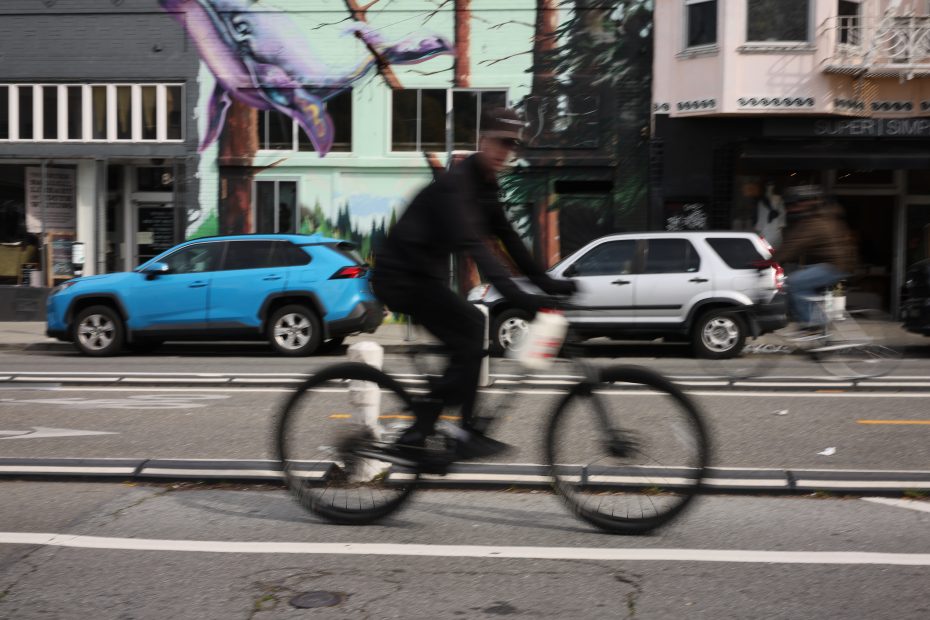 A person on a bicycle is in motion on a city street with a mural of a creature on a building and parked cars in the background.