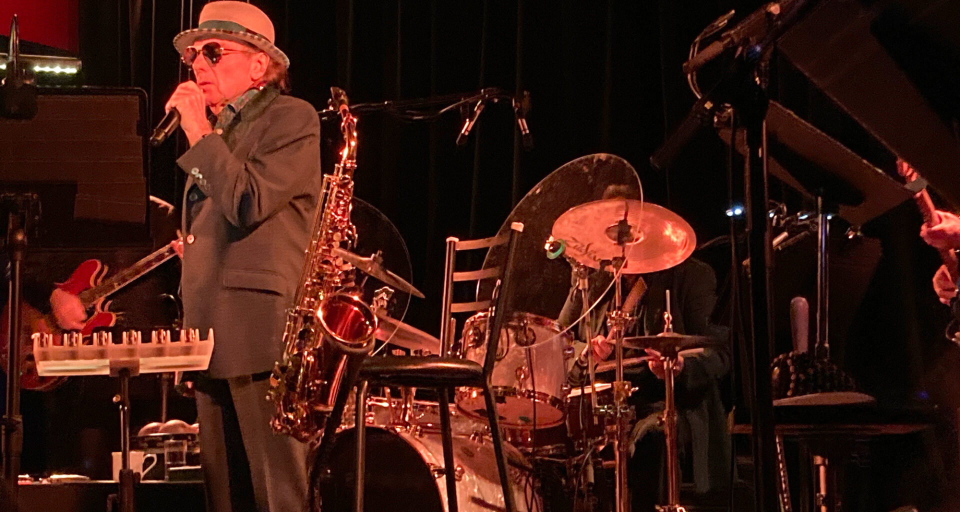 A man in a fedora hat and suit and sunglasses sings into a gold microphone on a stage, surrounded by instruments