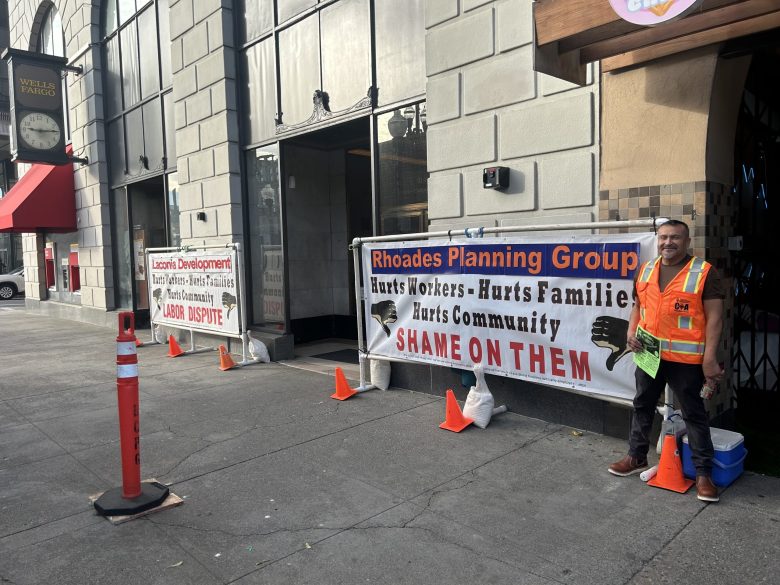 A worker in an orange vest stands next to a sign that reads, "Rhoades Planning Group hurts workers, hurts families hurts community. Shame on them"