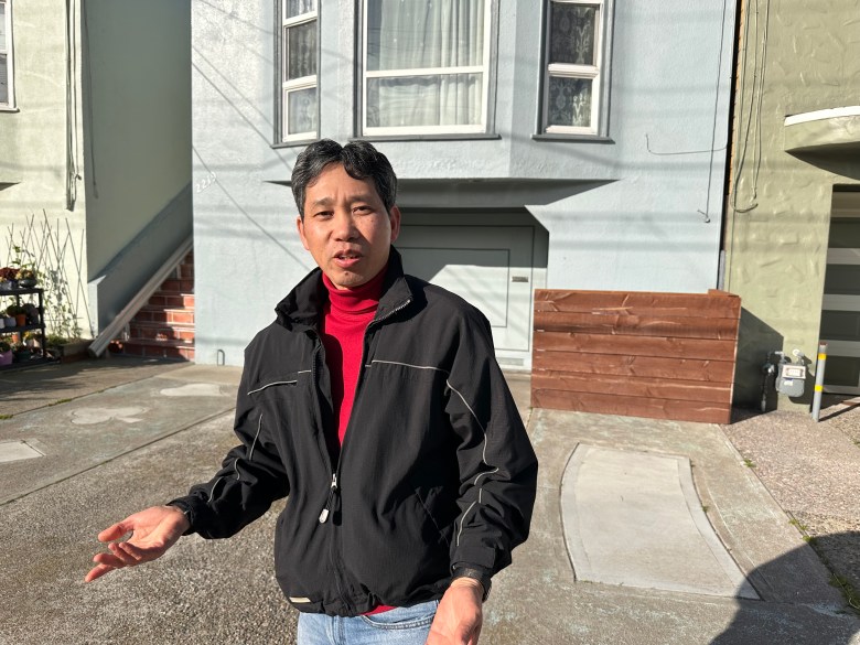 A man in a jacket stands on the street in a residential neighborhood facing the camera