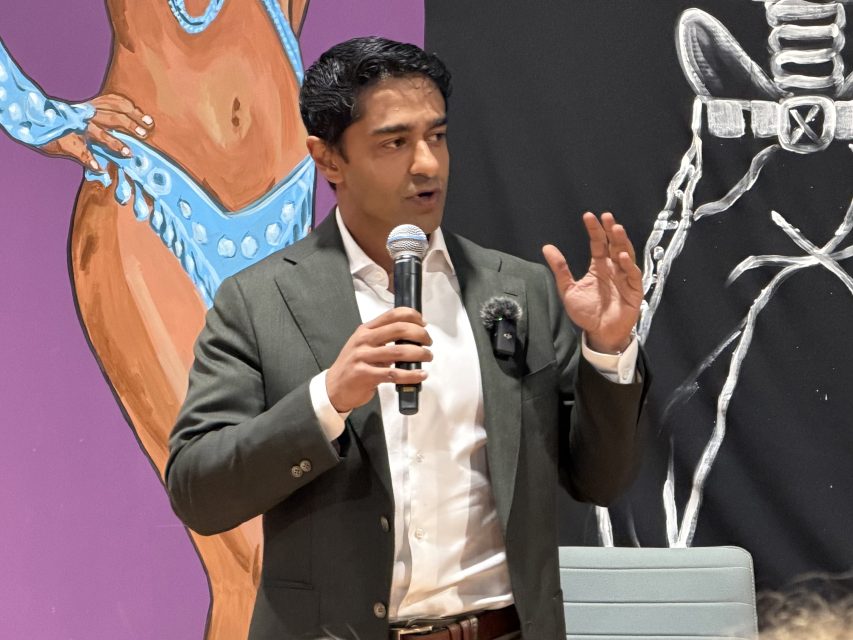 A man in a suit speaks into a microphone, gesturing with his hand, with colorful artwork featuring figures in the background.