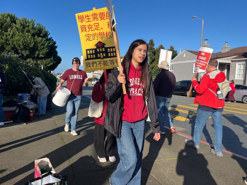 A group of people wearing red jackets hold signs and walk on a sidewalk during a school strike protest. One sign has Chinese text and an image of a school bus.