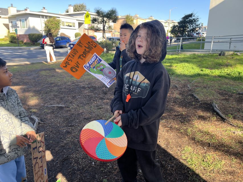 A child in a black hoodie holds a colorful paddle and stands outdoors with other kids. One child holds a sign that reads, "We stand by our teachers." Houses and trees are in the background.