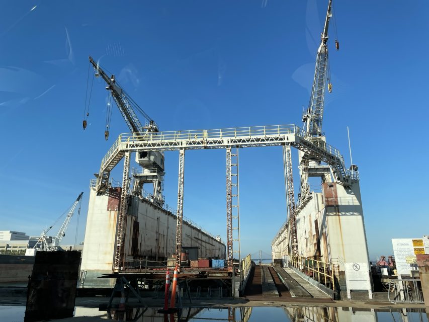 A large industrial dry dock with two cranes on either side, metal framework overhead, and a clear blue sky in the background.