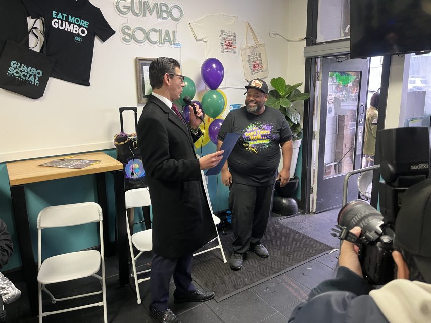 A man in a suit speaks into a microphone while holding a document, facing another man in casual clothing at an indoor event decorated with balloons. A photographer takes pictures nearby.