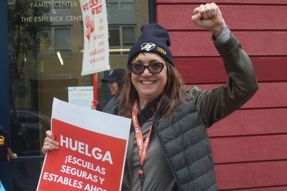 A woman wearing sunglasses and a beanie holds a "Huelga" sign and raises her fist outside a building, participating in a strike for safe and stable schools.