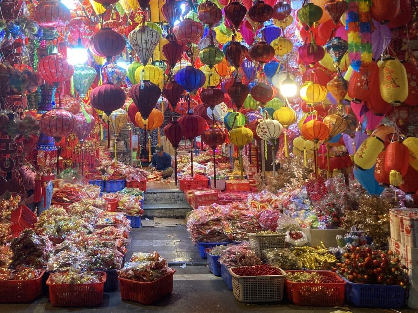 A market stall displays colorful paper lanterns hanging above baskets filled with assorted festive decorations and goods, with two people sitting in the background.