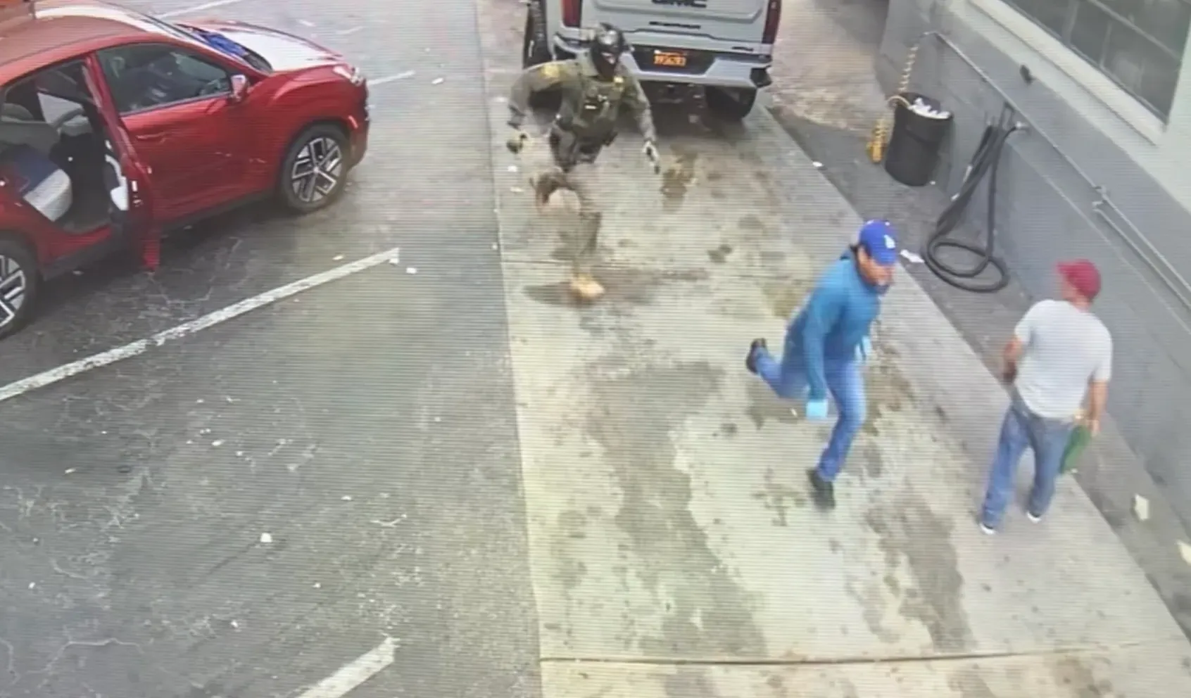 A man wearing blue clothing and a blue baseball cap runs from a masked man in body armor.