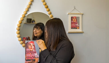Author Dr. Loretta Ramirez holding her book, 'The Wound & The Stitch,' while standing in front of a circular mirror and framed artwork.