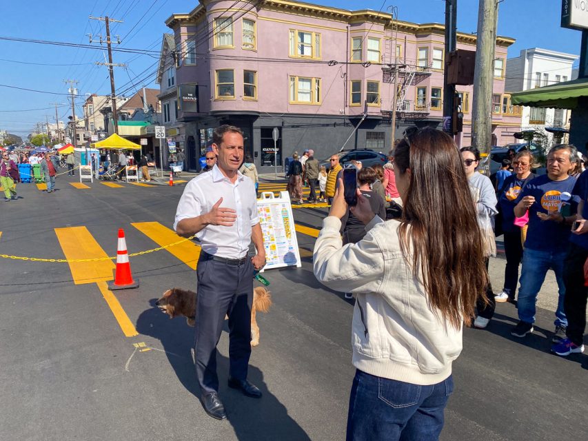 A man speaks while being recorded by a woman on a street with people, buildings, and tents in the background. There's a dog by his side and an orange traffic cone nearby.