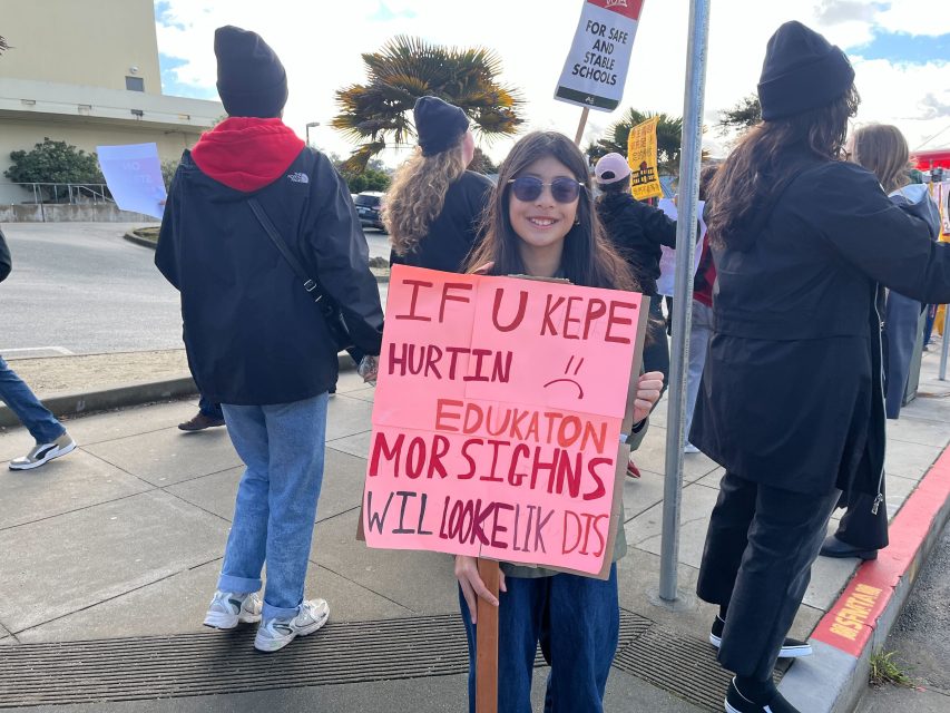 A person at a protest holds a sign with intentionally misspelled words about education, reading: "If u kepe hurtin edukaton mor signhs wil looke lik dis.