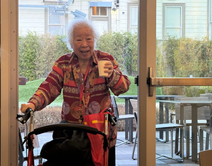 An elderly woman with a walker stands by a glass door, holding a cup and smiling. Outdoor seating and greenery are visible through the window.