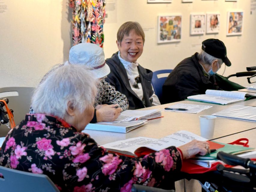 Four older adults sit at a table with books and papers, engaging in discussion in a brightly lit room with art displayed on the walls.