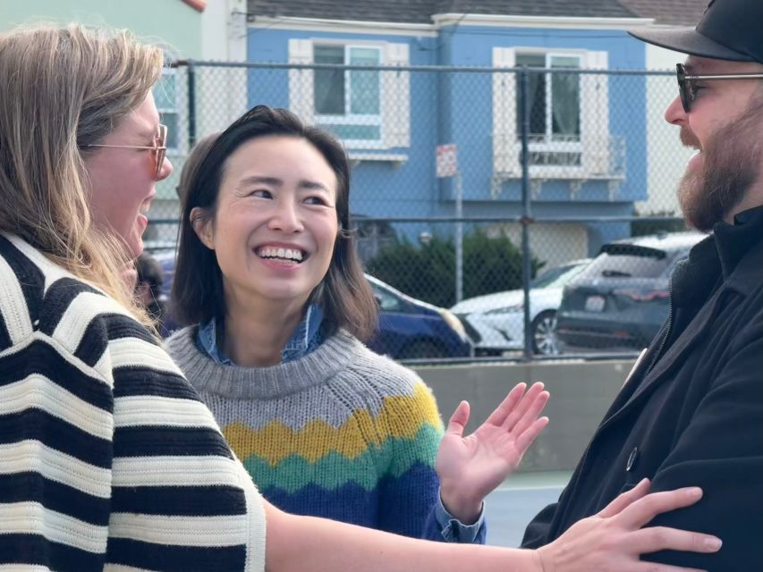 Three adults stand outdoors, talking and smiling near a fence with houses and parked cars in the background.