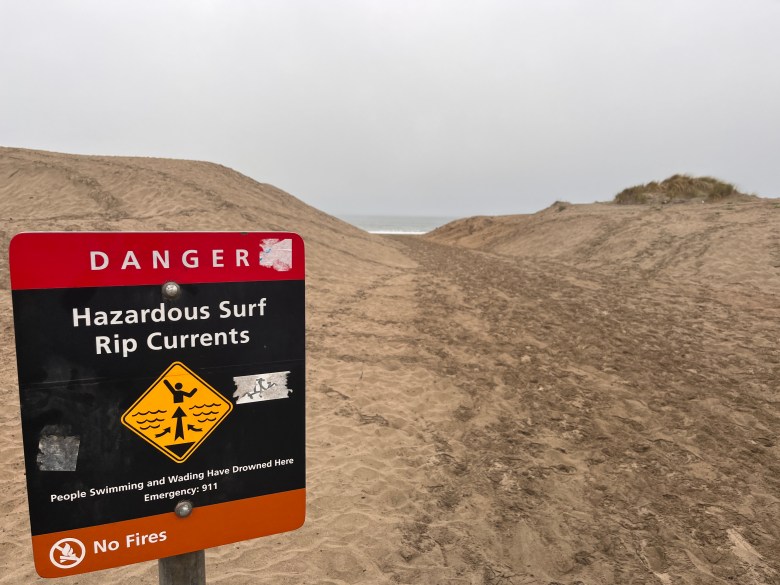 A sign warning of hazardous rip currents in front of sand dunes and a path that leads to Ocean Beach from the former Great Highway.