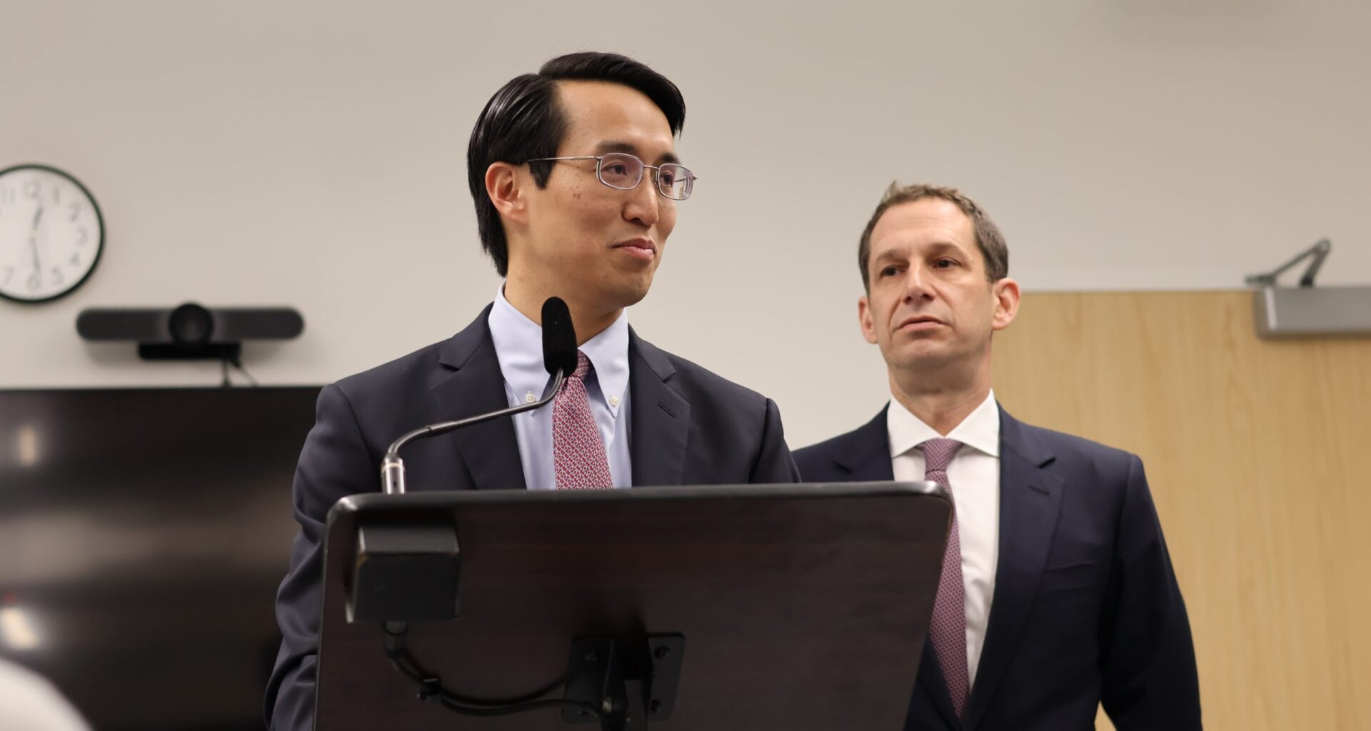 Two men in suits stand at a podium in an office setting. One speaks into a microphone while the other listens. A clock, television, and exit sign are visible in the background.