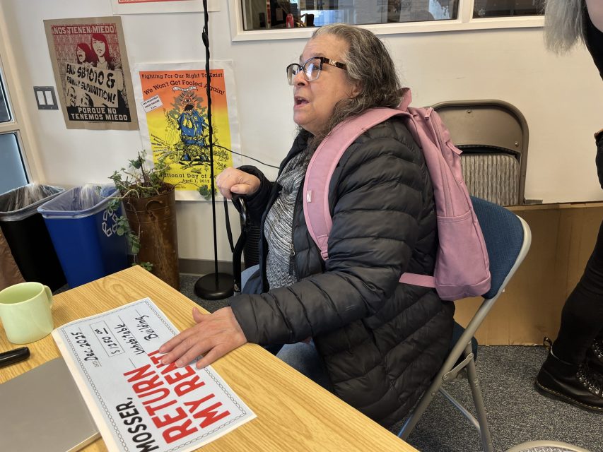 A woman with gray hair and glasses, wearing a pink backpack, sits at a table holding a cane and a "RETURN MY RENT" sign. Posters and a recycling bin are visible in the background.