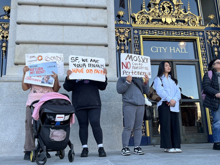 People stand in front of a city hall entrance holding protest signs about rent and tenants' rights; one person has a stroller, and ornate gold doors are visible behind them.