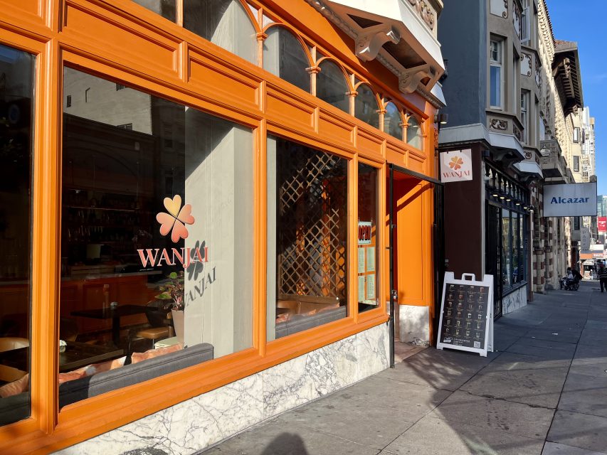 Street view of the exterior of Wanja restaurant with orange-framed windows and a marble base, next to Alcazar on a sunny day.