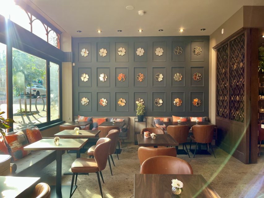 Sunlit café interior with brown leather chairs, wooden tables, and flower art on dark wall; large windows let in bright natural light.