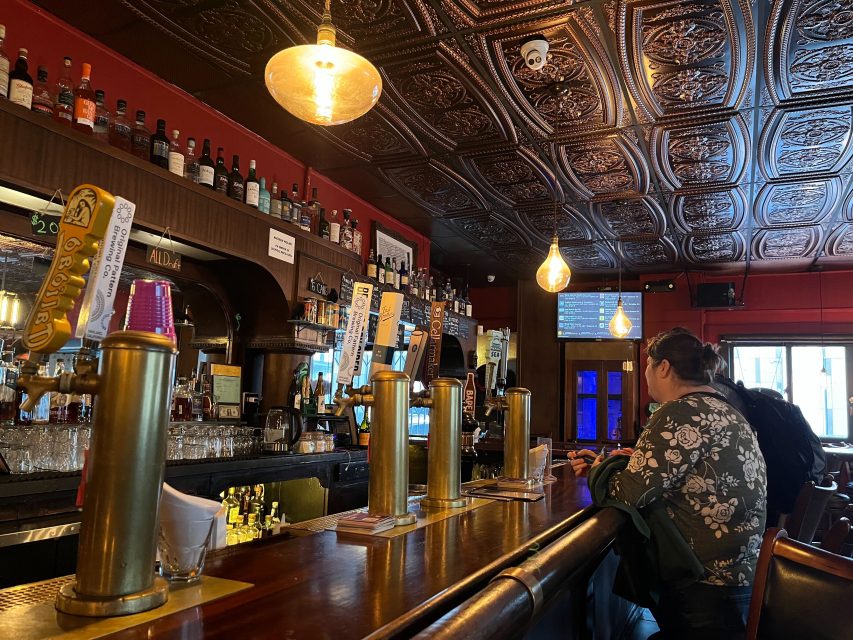A bar with ornate tin ceiling tiles, several beer taps, bottles on shelves, and two people sitting at the counter.