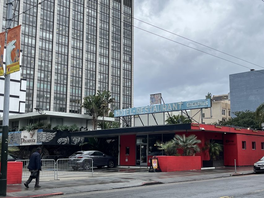 A red, single-story restaurant with a sign reading "Hotel Restaurant Cocktails" sits on a city street with tall buildings in the background and a person walking by in front.