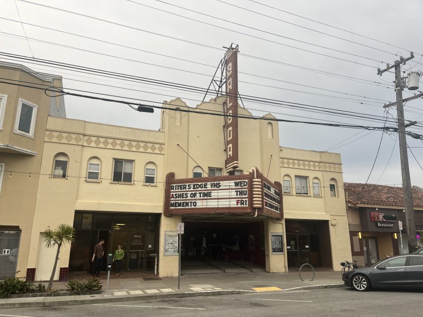 A beige art deco theater building with a marquee listing movies and showtimes, located on a city street with nearby shops and power lines overhead.