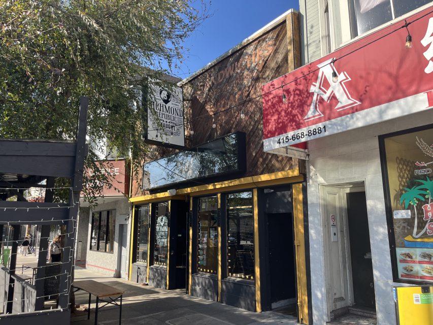 Street view of Richmond Republic Draught House bar with a black and white sign, next to a red storefront with a white "A" logo, on a sunny day.