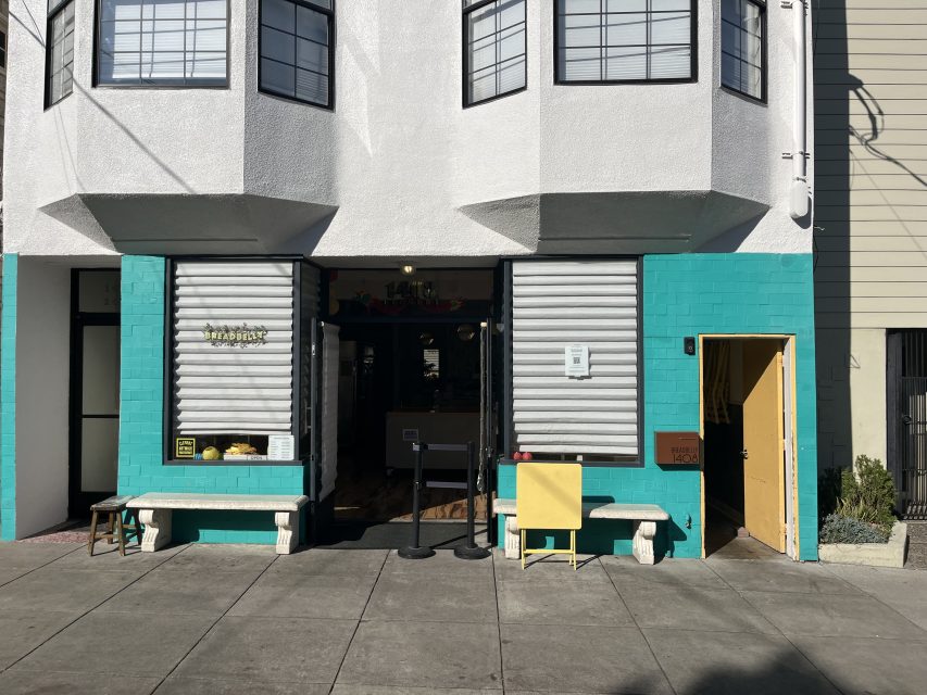 Street view of a turquoise and white storefront with two benches, a yellow door to the right, and partially closed metal shutters on the windows.