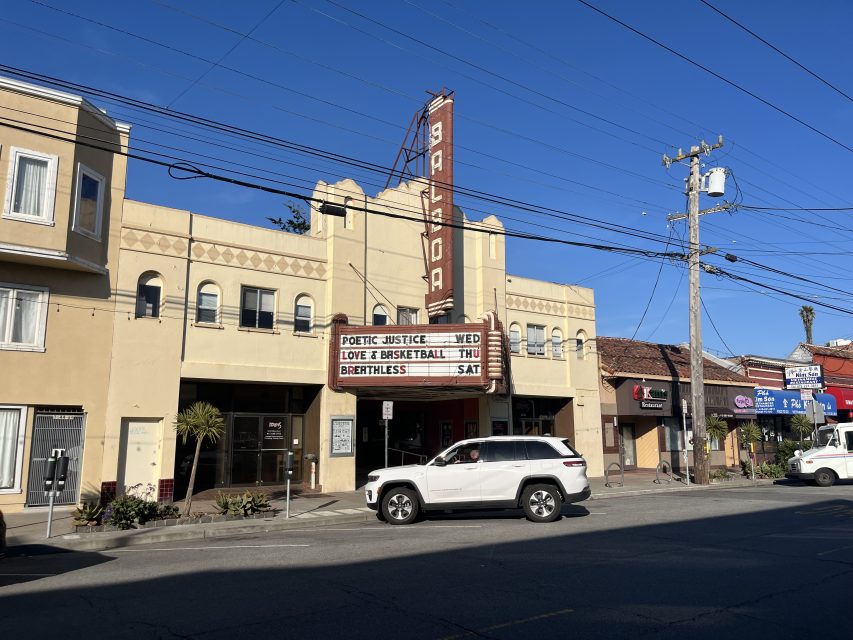 A beige Art Deco theater with a vertical “Balboa” sign displays movie titles on its marquee; a white SUV drives by on a sunny day.