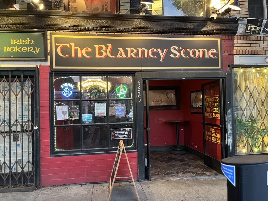Street view of The Blarney Stone pub entrance with neon signs in the window, a sandwich board outside, and neighboring Irish Bakery on the left.