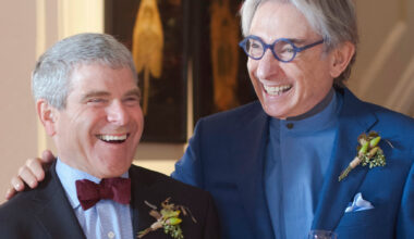 A man in a black suit and deep maroon bowtie smiles next to a man in blue suit and glasses