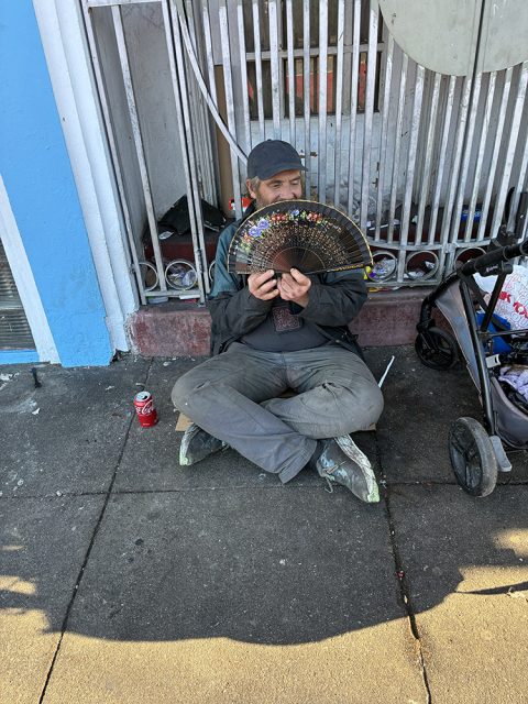 A person sits on a city sidewalk in front of a metal gate, holding a decorative fan. A stroller and a can of soda are nearby.