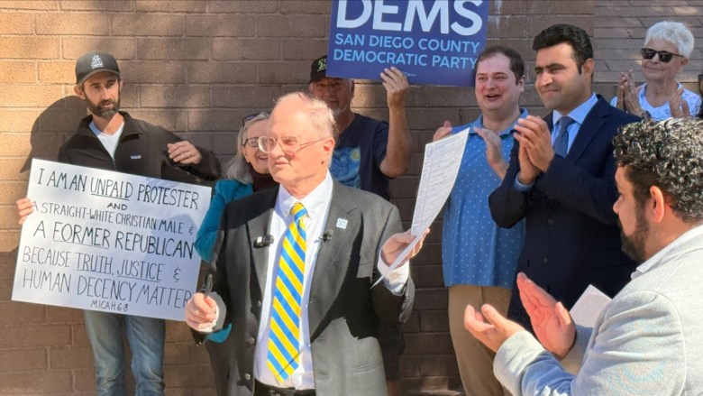 A group gathers in front of a brick wall; a man in a suit holds a document while others clap and hold various signs.