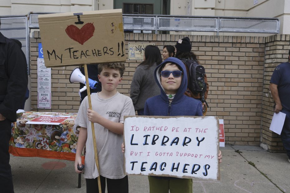 Two children stand outside holding protest signs in support of their teachers; one sign reads "I ❤️ My Teacher!!" and the other highlights backing teachers over library time.