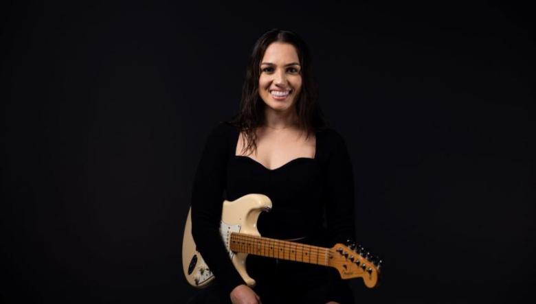 A woman dressed in black poses with a guitar against a black background