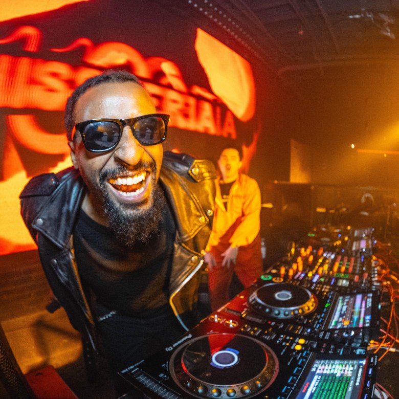 A man in sunglasses smiles while standing behind a DJ turntable setup