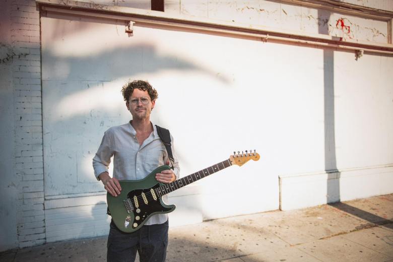 A man holding an electric guitar stands in front of a white wall outdoors