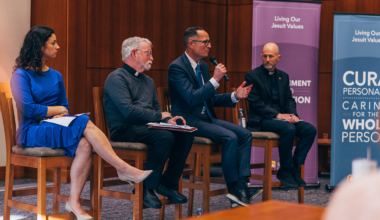 Four panelists sit in chairs with microphones in a lecture hall.