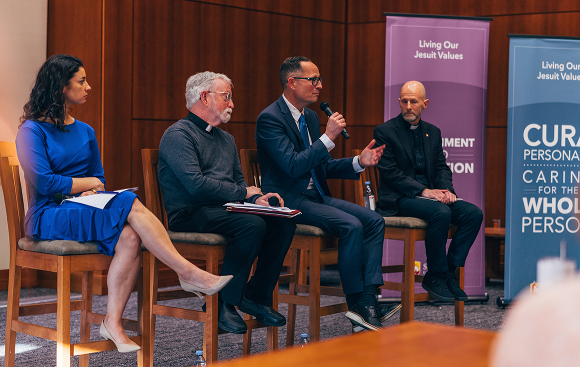 Four panelists sit in chairs with microphones in a lecture hall.