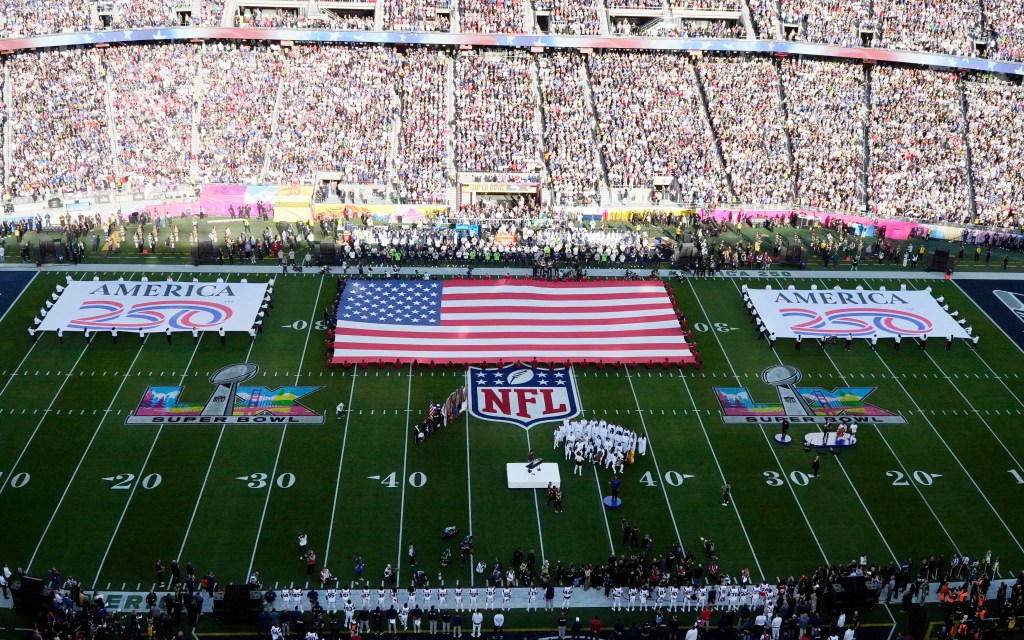 View of a Super Bowl football field with a large American flag spread across the 50-yard line, surrounded by cheerleaders and two large "America 250" banners, all under a stadium filled with spectators.
