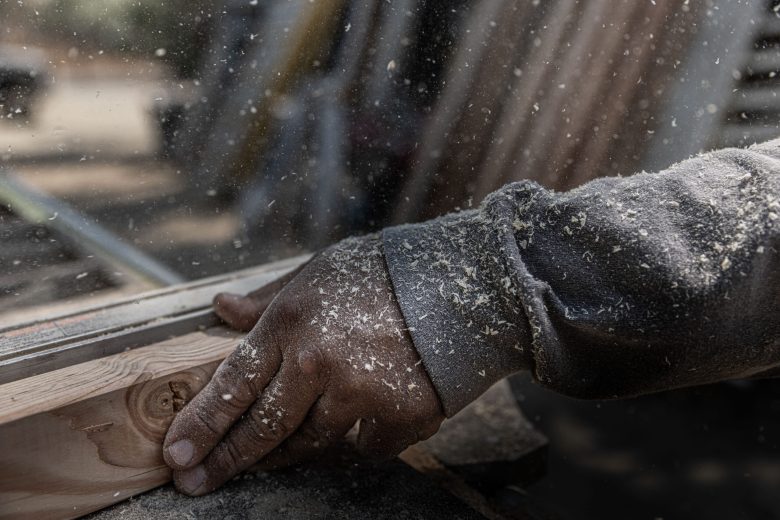 A hand covered in sawdust guides a wooden plank through a table saw. Sunlight filters through dust particles, creating a focused, industrious atmosphere.