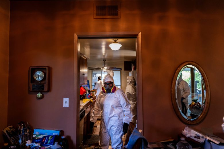 Person in a protective suit and respirator stands in a cluttered room. Others in similar attire work in the background. The mood is serious.