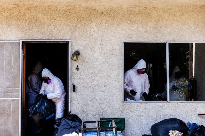 Two workers in hazmat suits and respirators clean a cluttered room. One is seen through an open door, the other through a window. The scene feels tense.