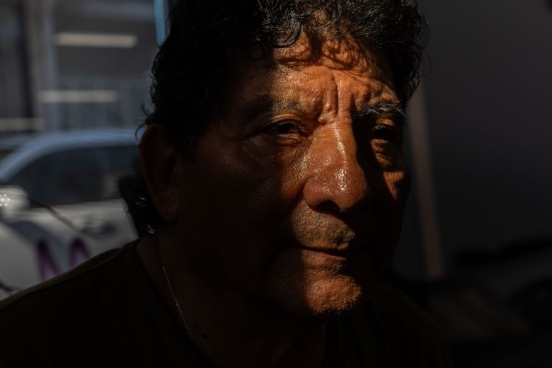 Elderly man in partial shadow and sunlight, revealing deep lines and a thoughtful expression, suggesting contemplation. Urban background blurred.