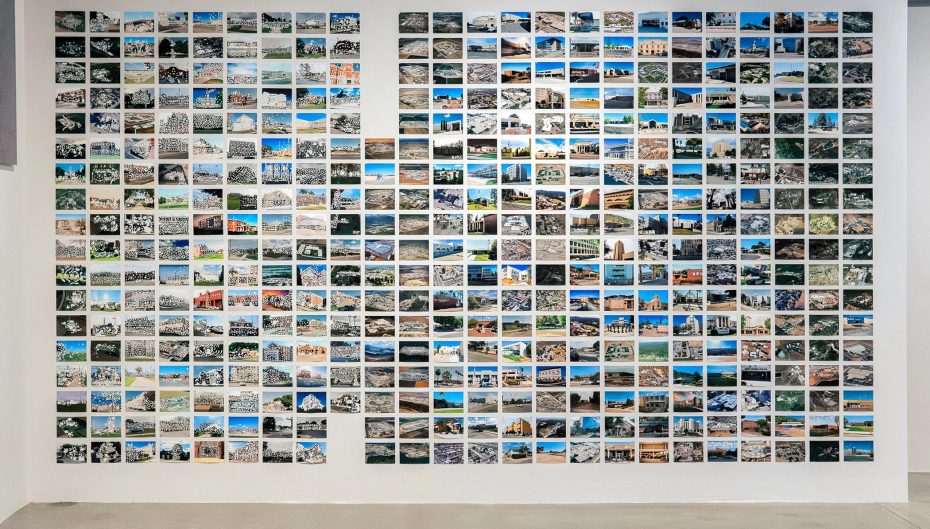 A large grid of small photographs is displayed on a white wall, each depicting various landscapes, buildings, and scenes from different locations, reminiscent of collections often found in museums.