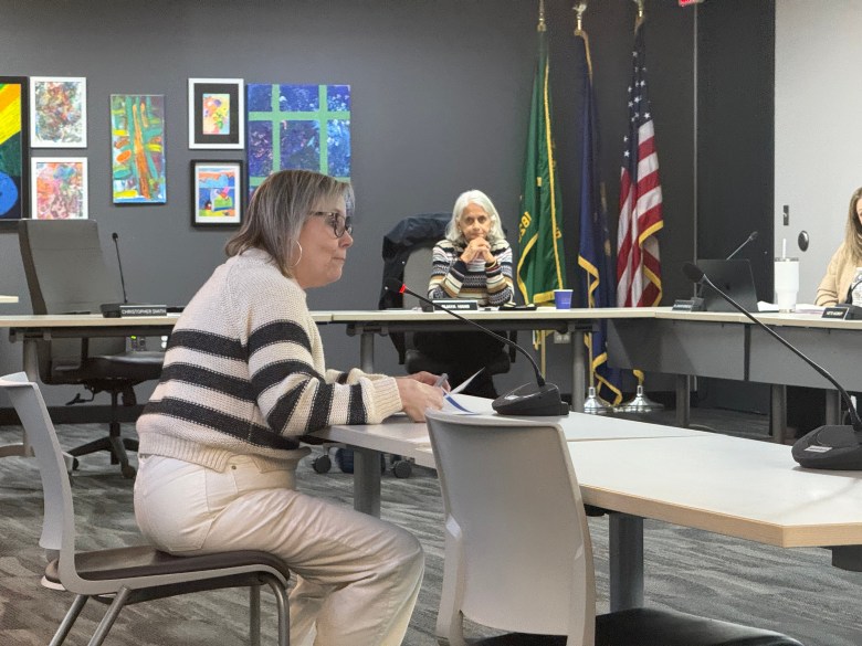 A woman sits at a table speaking at a public event as another woman looks on