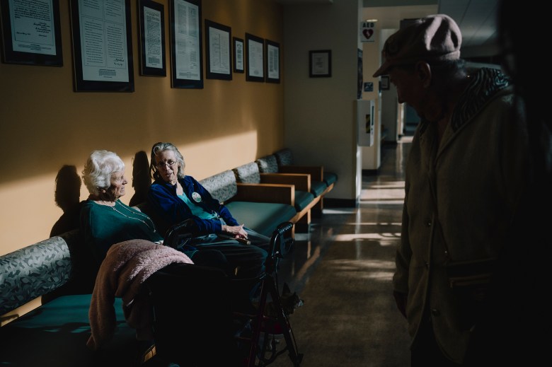 Two older adults sit on a bench along a hallway wall displaying framed documents, lit by a patch of sunlight, while another older adult wearing a cap stands nearby facing them.