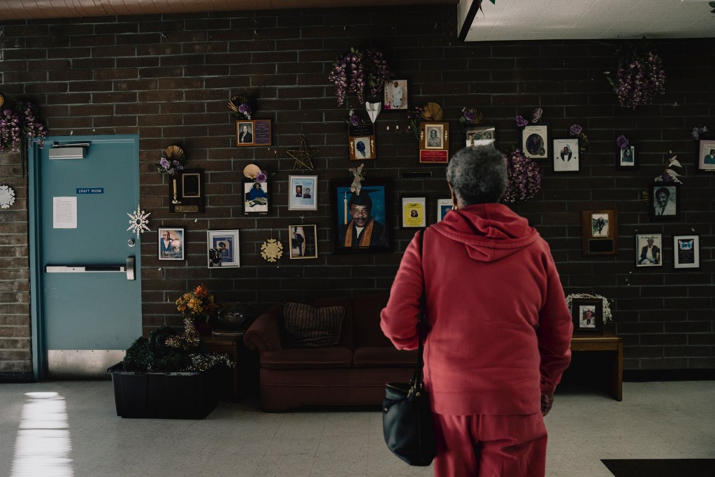 A person in a red outfit stands facing a brick wall covered with framed photos, decorations, and flowers, with a couch and a door labeled “Craft Room” nearby.